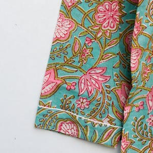 Op de afbeelding: Close-up van een turquoise stof met een bloemenpatroon in roze en groen. Het ontwerp heeft grote bloemen en kronkelende wijnranken. De randen zijn afgewerkt met een witte rand. De stof lijkt katoen te zijn.