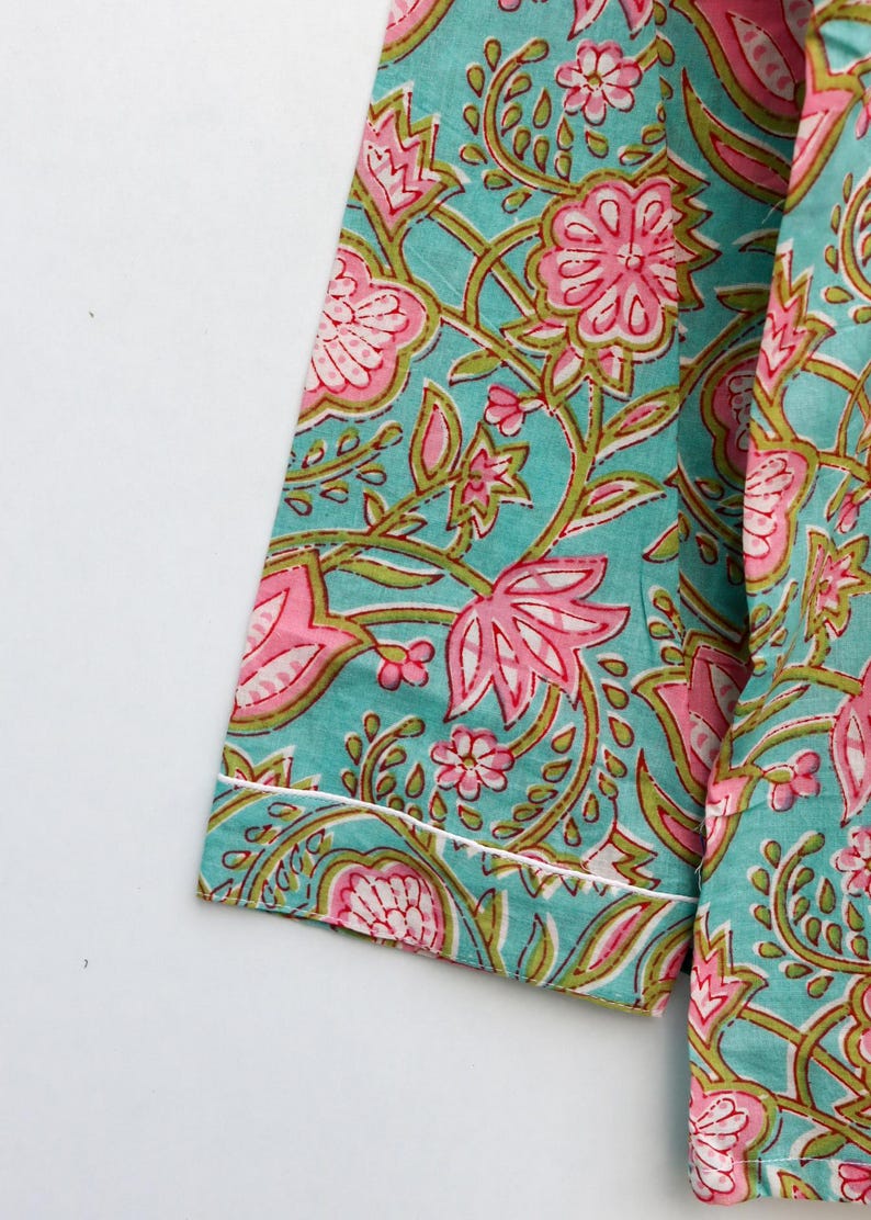Op de afbeelding: Close-up van een turquoise stof met een bloemenpatroon in roze en groen. Het ontwerp heeft grote bloemen en kronkelende wijnranken. De randen zijn afgewerkt met een witte rand. De stof lijkt katoen te zijn.