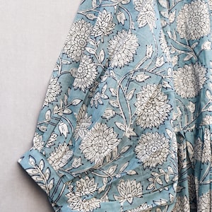 May include: Close-up of a light blue fabric with a white floral pattern. The design features large, stylized flowers and leaves. The fabric appears to be part of a garment, with a sleeve and cuff visible.