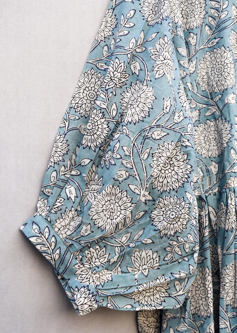 May include: Close-up of a light blue fabric with a white floral pattern. The design features large, stylized flowers and leaves. The fabric appears to be part of a garment, with a sleeve and cuff visible.
