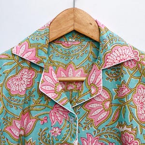 Op de afbeelding: Een button-down pyjamajas met een bloemenpatroon. De stof is licht turquoise met roze en groene bloemmotieven. De kraag en manchetten zijn afgewerkt met witte biezen. De top hangt aan een houten hanger.