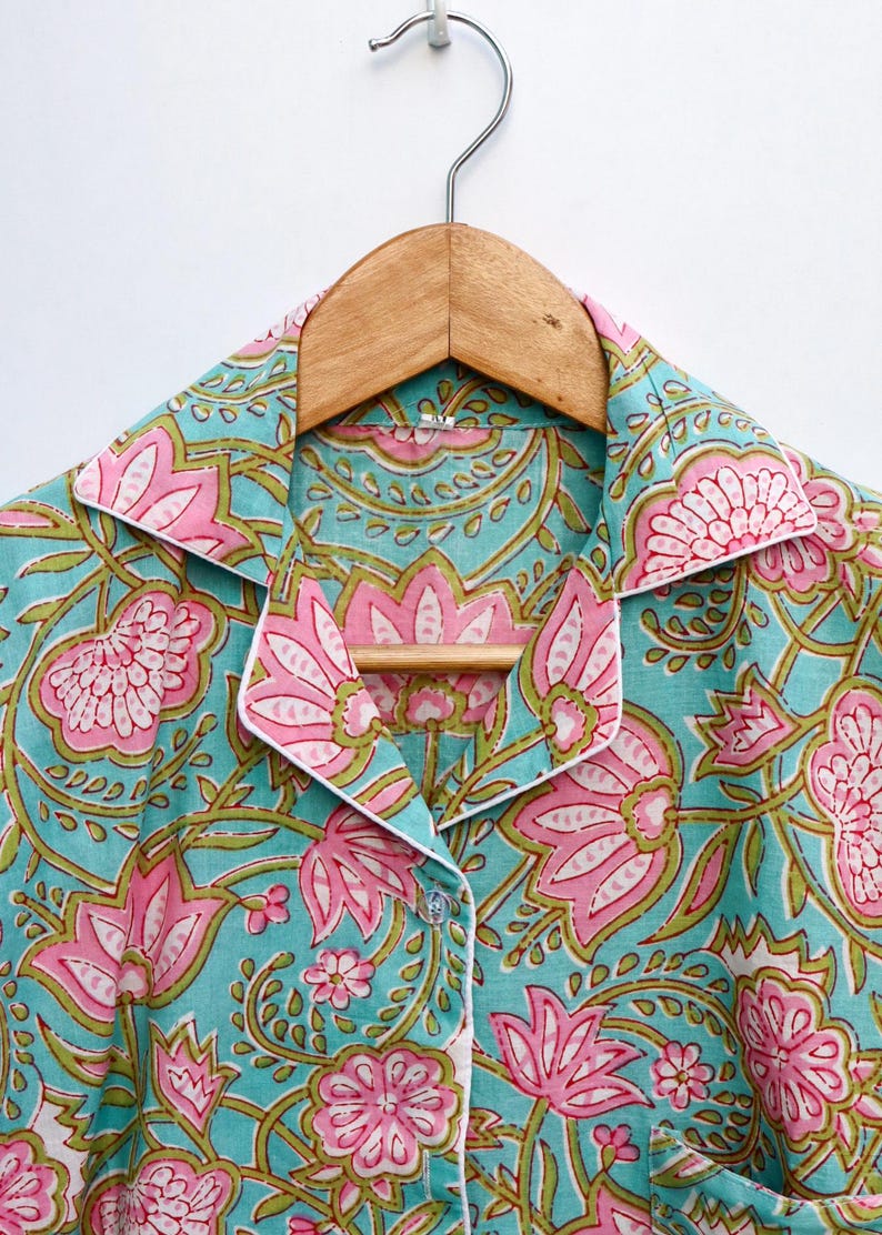 Op de afbeelding: Een button-down pyjamajas met een bloemenpatroon. De stof is licht turquoise met roze en groene bloemmotieven. De kraag en manchetten zijn afgewerkt met witte biezen. De top hangt aan een houten hanger.