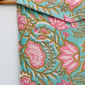 Op de afbeelding: Een turquoise stoffen etui met een roze en groen bloemenpatroon, hangend aan een houten hanger. Het ontwerp toont grote roze bloemen en groene bladeren. De etui is waarschijnlijk voor opslag.