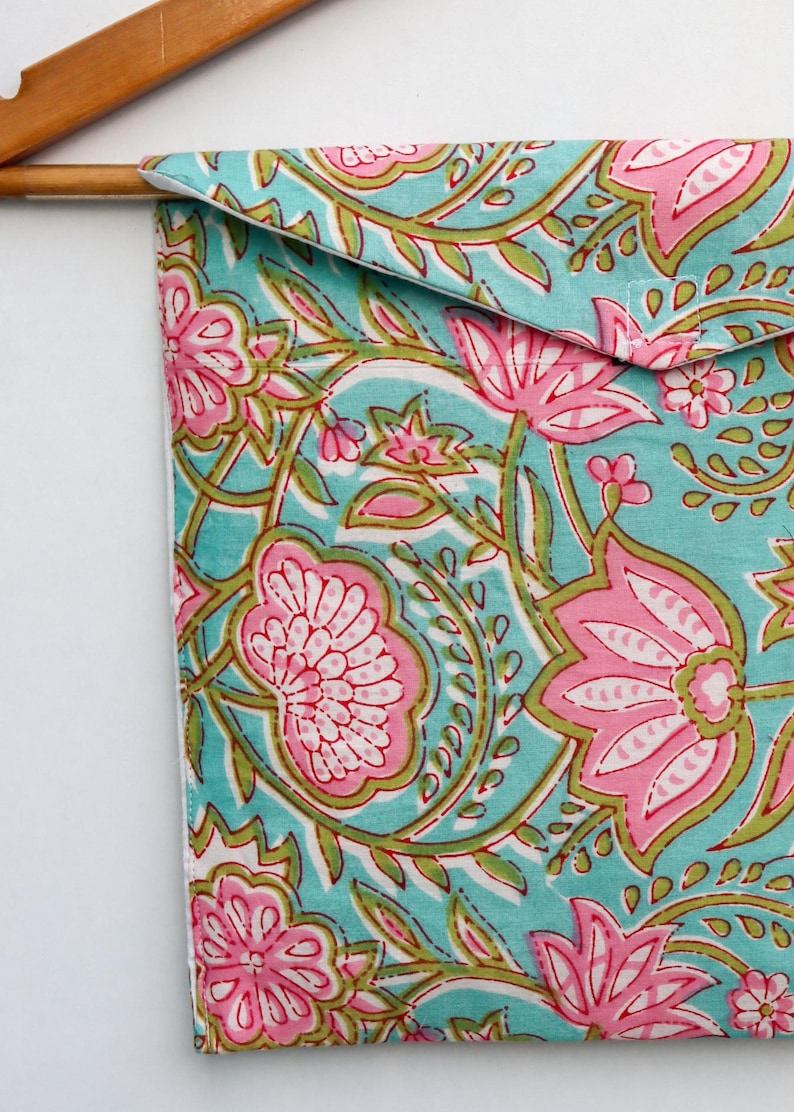 Op de afbeelding: Een turquoise stoffen etui met een roze en groen bloemenpatroon, hangend aan een houten hanger. Het ontwerp toont grote roze bloemen en groene bladeren. De etui is waarschijnlijk voor opslag.