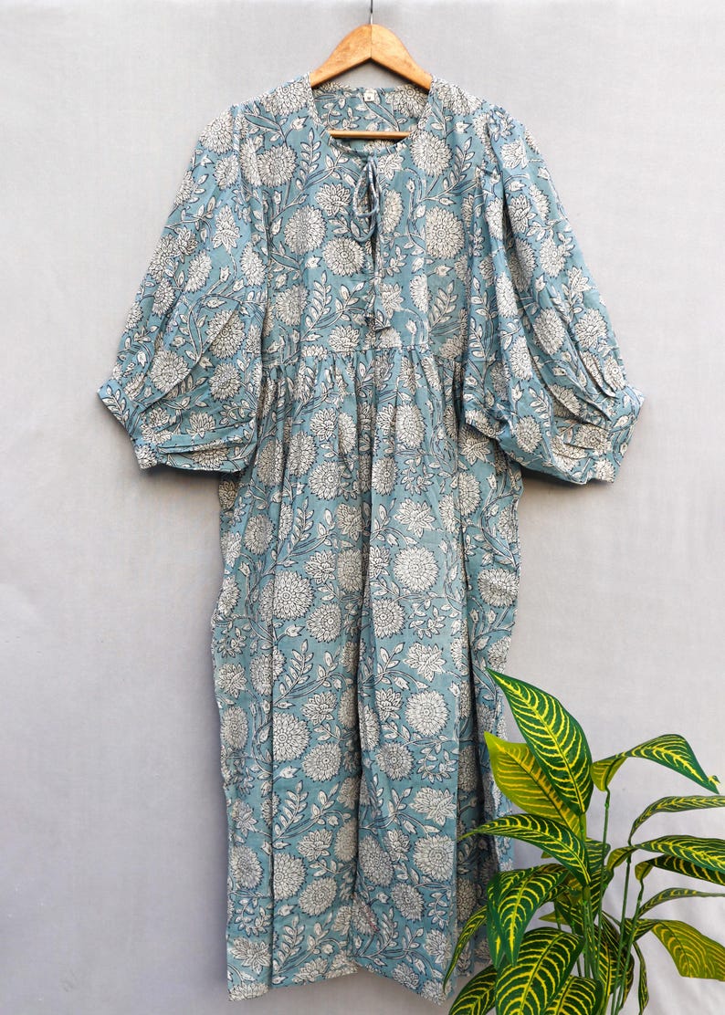 May include: A light blue dress with a white floral pattern, hanging on a wooden hanger. The dress features a round neckline with a tie, three-quarter sleeves, and a gathered waist. The fabric has a soft, flowing appearance, suitable for casual wear.