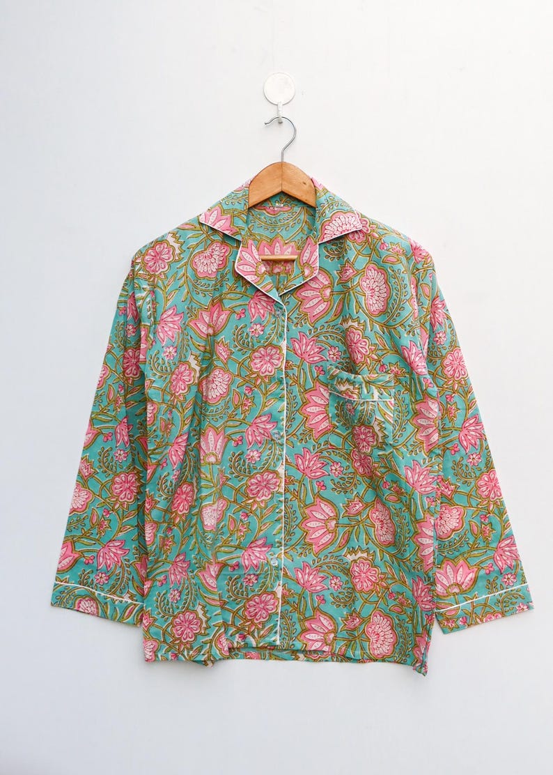 Op de afbeelding: Een pyjamajas met lange mouwen en een bloemenpatroon in teal, roze en groen. De top heeft een kraag, een borstzak en witte biezen. Het kledingstuk hangt aan een houten hanger tegen een witte achtergrond.