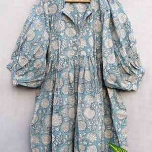 May include: A light blue dress with a white floral pattern. The dress has a collared neckline, long sleeves with gathered cuffs, and a gathered waist. The fabric appears to be cotton or a similar lightweight material. The dress is hanging on a wooden hanger.