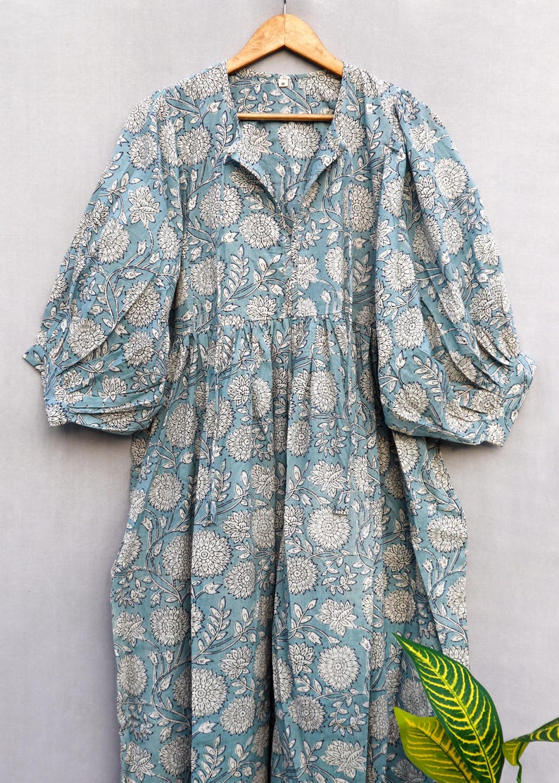 May include: A light blue dress with a white floral pattern. The dress has a collared neckline, long sleeves with gathered cuffs, and a gathered waist. The fabric appears to be cotton or a similar lightweight material. The dress is hanging on a wooden hanger.