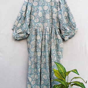 May include: A light blue dress with a white floral pattern. The dress has long, gathered sleeves and a gathered waist. It is hanging on a wooden hanger against a light gray background. The dress is made of a lightweight fabric.