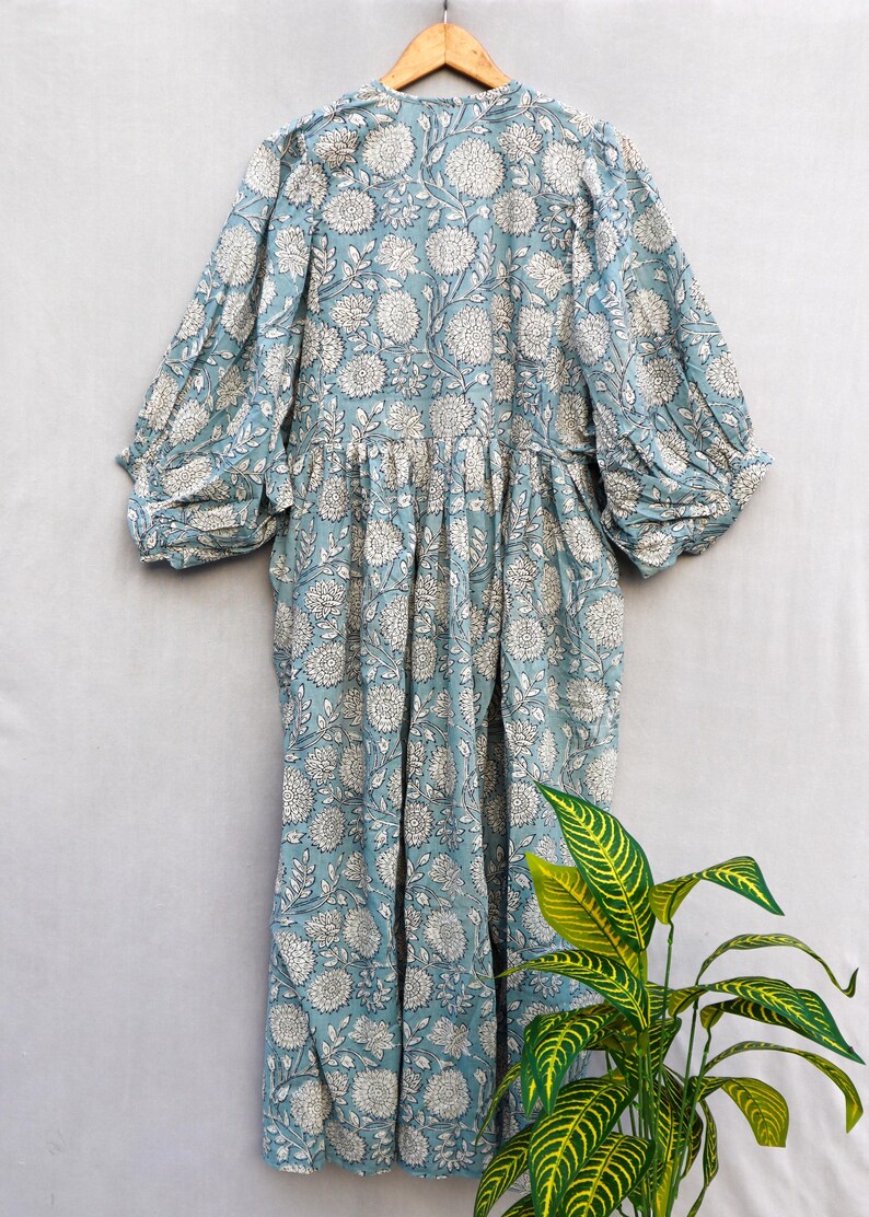 May include: A light blue dress with a white floral pattern. The dress has long, gathered sleeves and a gathered waist. It is hanging on a wooden hanger against a light gray background. The dress is made of a lightweight fabric.