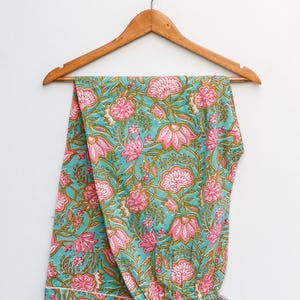 Op de afbeelding: Turquoise pyjamabroek met bloemenprint in roze en groen, hangend aan een houten hanger. Het bloemenpatroon heeft grote roze bloemen en groene bladeren. De broek is gemaakt van zachte stof.