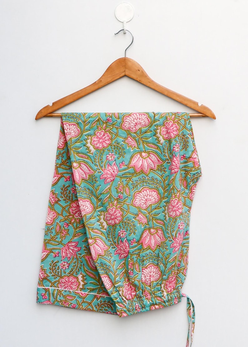Op de afbeelding: Turquoise pyjamabroek met bloemenprint in roze en groen, hangend aan een houten hanger. Het bloemenpatroon heeft grote roze bloemen en groene bladeren. De broek is gemaakt van zachte stof.