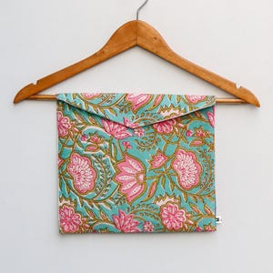 Op de afbeelding: Een turquoise etui met een roze bloemenpatroon, hangend aan een houten kleerhanger. Het ontwerp toont herhaalde bloemen en bladeren in roze en groen. De etui heeft een rechthoekige vorm en een klepsluiting.