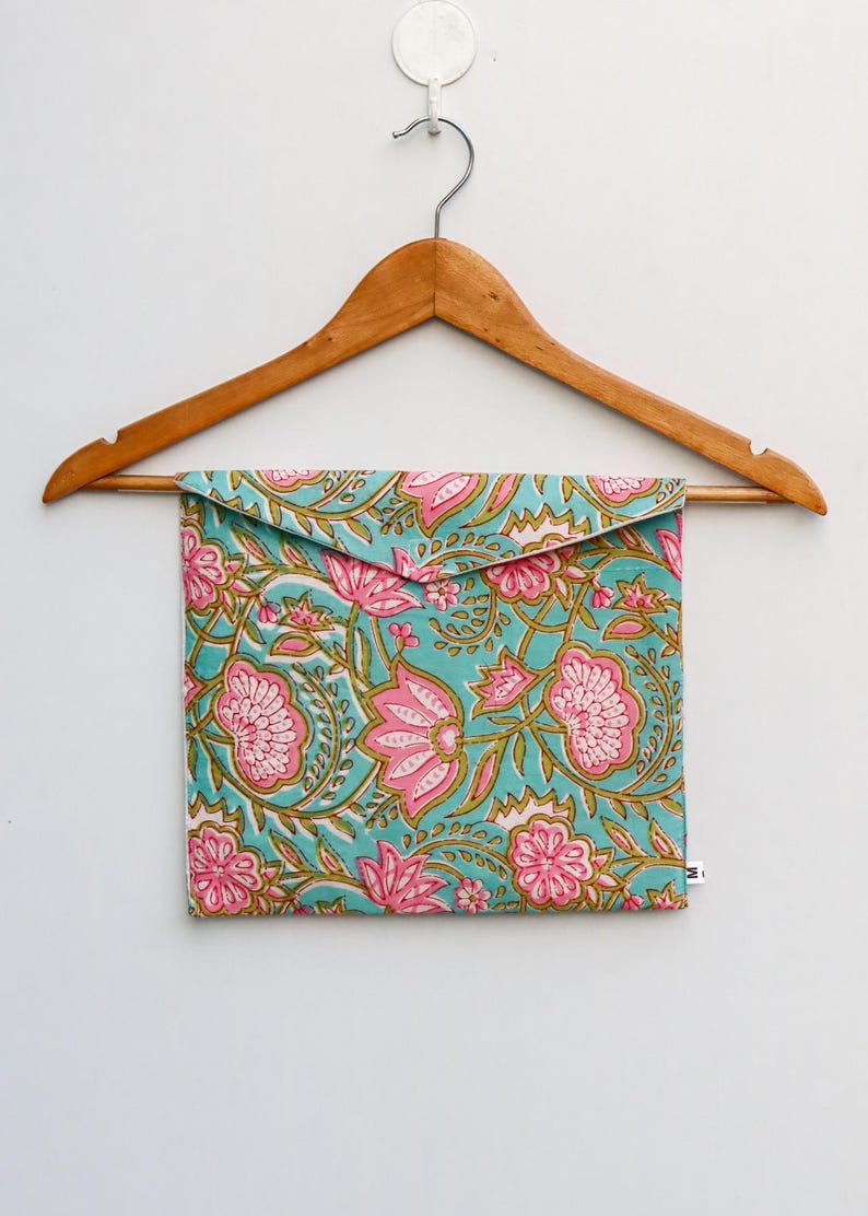 Op de afbeelding: Een turquoise etui met een roze bloemenpatroon, hangend aan een houten kleerhanger. Het ontwerp toont herhaalde bloemen en bladeren in roze en groen. De etui heeft een rechthoekige vorm en een klepsluiting.