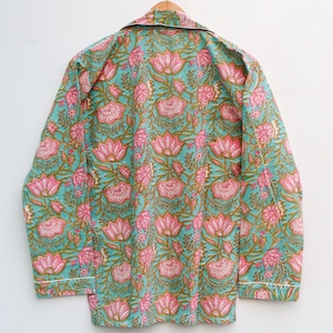 Op de afbeelding: Een turquoise pyjamajas met een roze bloemenpatroon en witte bies. De top met lange mouwen hangt aan een houten hanger. Het bloemmotief heeft grote roze lotusbloemen en groene bladeren.