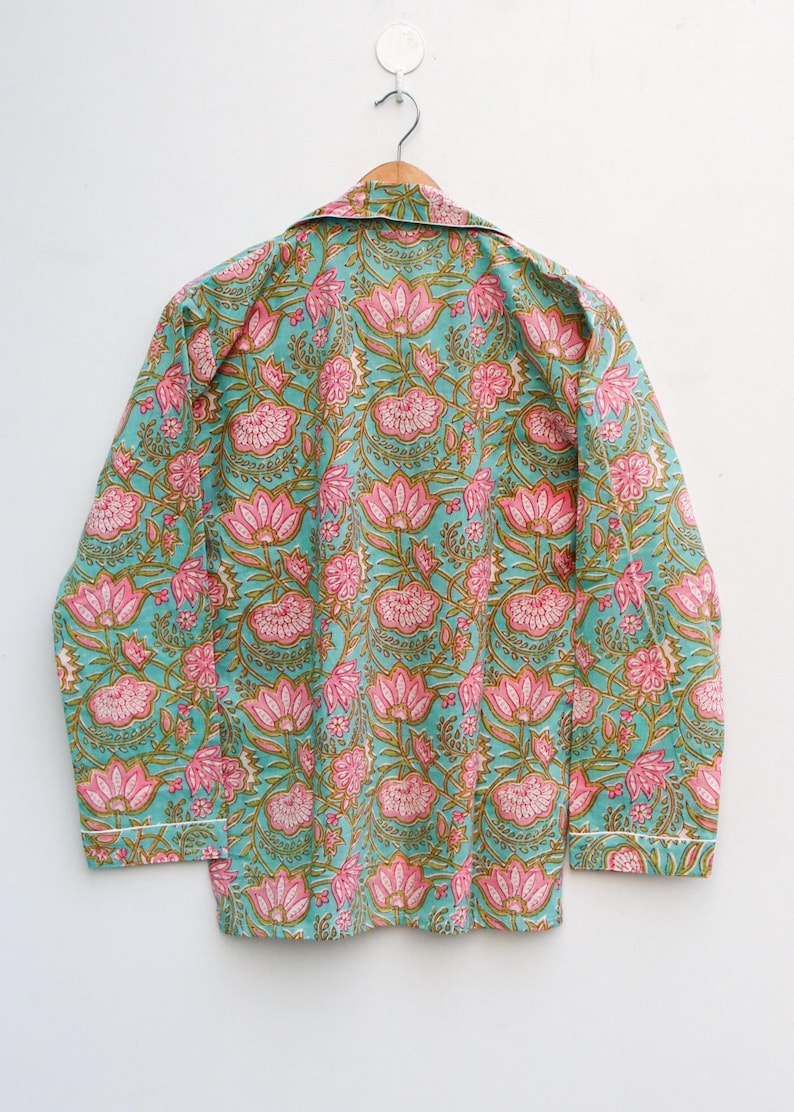 Op de afbeelding: Een turquoise pyjamajas met een roze bloemenpatroon en witte bies. De top met lange mouwen hangt aan een houten hanger. Het bloemmotief heeft grote roze lotusbloemen en groene bladeren.