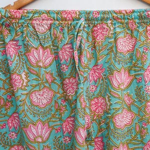 Op de afbeelding: Close-up van een pyjamabroek met een turquoise achtergrond en een roze bloemenpatroon. De broek heeft een taille met trekkoord en hangt aan een houten hanger. Het bloemmotief omvat grote lotusachtige bloemen en kleinere bloesems.