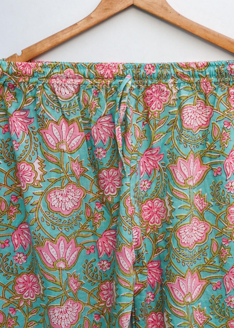 Op de afbeelding: Close-up van een pyjamabroek met een turquoise achtergrond en een roze bloemenpatroon. De broek heeft een taille met trekkoord en hangt aan een houten hanger. Het bloemmotief omvat grote lotusachtige bloemen en kleinere bloesems.