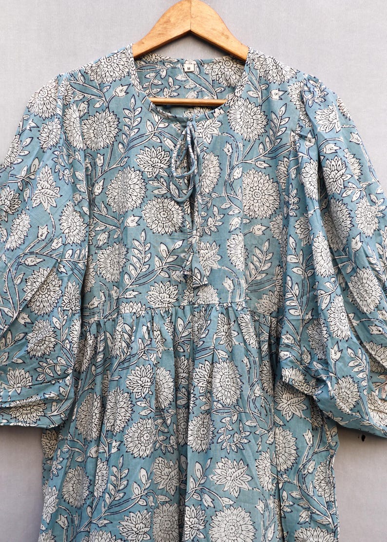 May include: A light blue dress with a white floral pattern. The dress has long sleeves, a round neckline with a tie, and a tiered skirt. The fabric appears to be lightweight and suitable for warm weather.