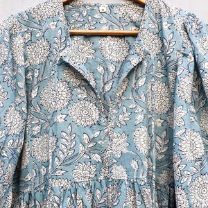 May include: A light blue dress with a white floral pattern. The dress is hanging on a wooden hanger. The floral design includes large flowers and leaves. The dress has a collared neckline and long sleeves.