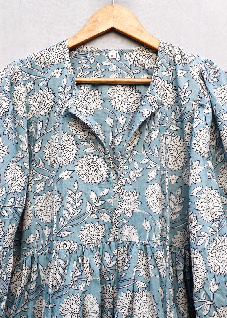 May include: A light blue dress with a white floral pattern. The dress is hanging on a wooden hanger. The floral design includes large flowers and leaves. The dress has a collared neckline and long sleeves.