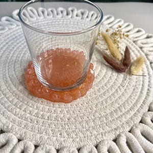 Può includere: Un bicchiere di vetro trasparente è appoggiato su un sottobicchiere in resina color pesca con scaglie d'oro. Il sottobicchiere ha un bordo smerlato. Il sottobicchiere e il bicchiere sono su un sottopiatti bianco intrecciato. Fiori secchi a destra.