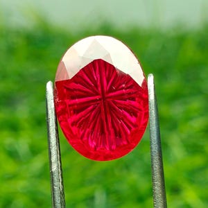 May include: An oval-shaped, vibrant red gemstone featuring a sunburst-style faceted pattern on its surface. The gemstone is held securely by a pair of silver-toned metal tweezers, showcasing its translucent quality and deep crimson color.
