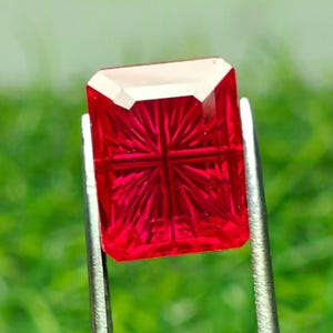 May include: A vibrant red gemstone with a rectangular shape and a precision-cut sunburst pattern on its surface. The stone is held by a pair of silver-toned metal tweezers, showcasing its translucent quality and deep crimson color throughout.