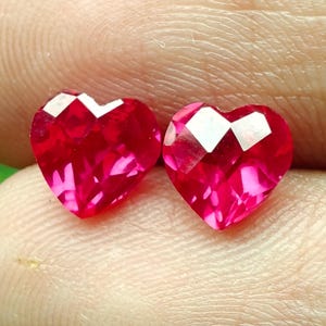 Puede incluir: Dos piedras preciosas rojas en forma de corazón con un corte facetado que refleja la luz. Estas pequeñas piedras pulidas exhiben un tono carmesí intenso y una silueta de corazón simétrica, mostrando facetas geométricas precisas.