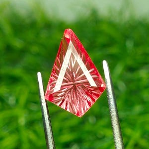 May include: A kite-shaped, faceted pink gemstone held by metal tweezers. The stone features a complex, star-like geometric pattern cut into its surface, creating sharp, radiating lines that catch the light across its translucent, vibrant pink body.