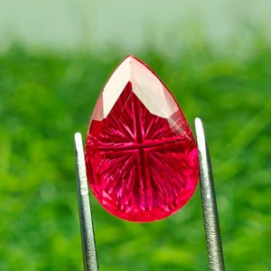 Puede incluir: Una gema roja vibrante en forma de pera con un detallado tallado radial en forma de estrella en su superficie. La piedra está sujeta por unas pinzas de metal plateado, mostrando su calidad translúcida y su intrincado patrón facetado simétrico.