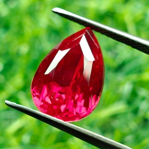 Puede incluir: Una gema roja vibrante en forma de pera sostenida por unas pinzas de metal oscuro. La piedra presenta una superficie facetada que refleja la luz, creando brillos intensos en su cuerpo carmesí profundo, mostrando un aspecto claro y pulido.