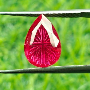 Peut inclure: Une pierre précieuse en forme de goutte d'eau, de couleur rouge vif, tenue par une pince argentée sur un fond vert flou. La gemme présente une facette blanche en forme de V en haut et des coupes internes complexes.