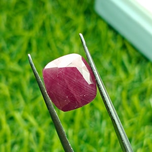 Pode incluir: Uma gema de rubi facetada com corte almofada, segurada por pinças. O rubi é de cor vermelha profunda e rica, com uma superfície polida. O fundo é verde desfocado, sugerindo um ambiente natural.