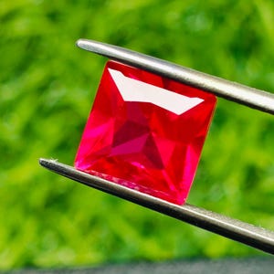 Puede incluir: Un rubí rectangular facetado de color rojo brillante es sujetado por unas pinzas plateadas sobre un fondo verde difuminado. El color rojo intenso y los cortes precisos de la gema crean un brillo que resalta su claridad.