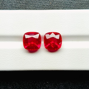 May include: Two vibrant red cushion-cut gemstones. The gems are displayed on a white surface with a textured pattern. The stones have a faceted design, reflecting light. The background is a dark gray color.