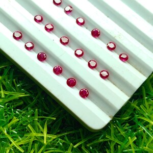 May include: A collection of small, round, faceted, deep red gemstones arranged on a white, ridged tray. The gemstones are evenly spaced and appear to be rubies. The tray is set against a background of bright green artificial grass.