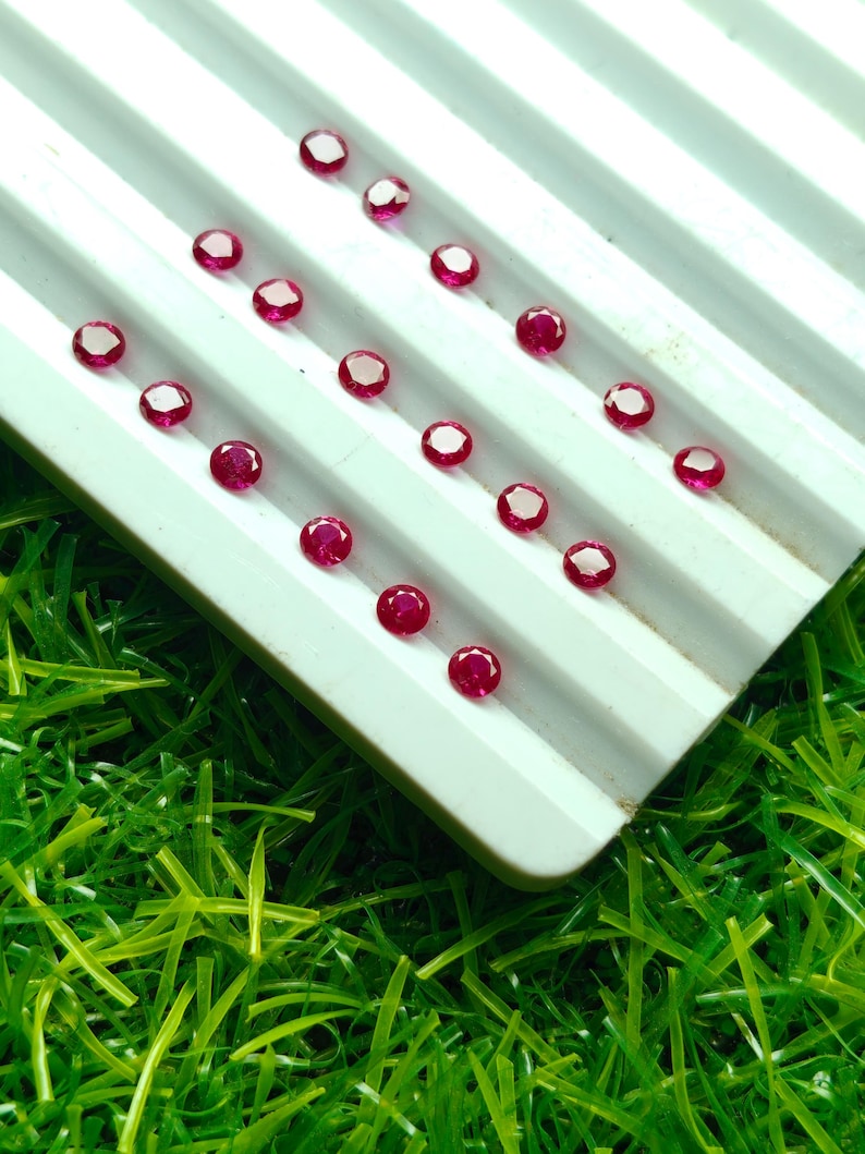 May include: A collection of small, round, faceted, deep red gemstones arranged on a white, ridged tray. The gemstones are evenly spaced and appear to be rubies. The tray is set against a background of bright green artificial grass.