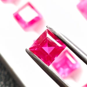Könnte beinhalten: Ein quadratisch geschliffener, leuchtend rosa Edelstein wird von einer Pinzette gehalten, die seine Facetten zeigt. Im Hintergrund sind weitere ähnliche Edelsteine zu sehen, was auf eine Sammlung oder Ausstellung von Edelsteinen hindeutet. Die leuchtend rosa Farbe hebt sich vom weißen Hintergrund ab.
