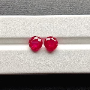 May include: Two faceted, oval-cut, bright red gemstones. The gems are displayed on a white surface with a textured pattern, set against a gray background. The stones are likely rubies, showcasing a vibrant color and clarity.