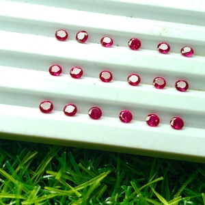 May include: A collection of small, round, faceted rubies arranged on a white surface. The gemstones are a deep red color with a bright, reflective surface. The rubies are displayed on a white tray against a backdrop of green grass.
