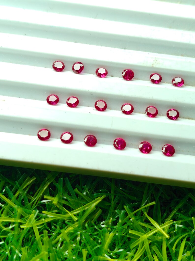 May include: A collection of small, round, faceted rubies arranged on a white surface. The gemstones are a deep red color with a bright, reflective surface. The rubies are displayed on a white tray against a backdrop of green grass.