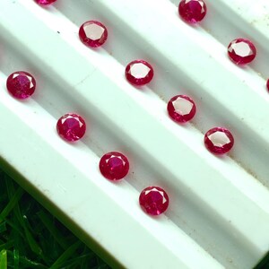 May include: A collection of round, faceted, deep red gemstones, possibly rubies, arranged on a white, ridged surface. The gems have a bright, reflective quality, and are set against a backdrop of green.