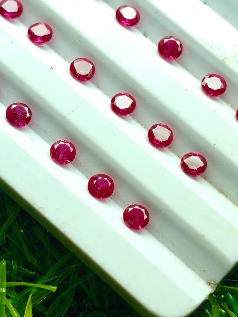 May include: A collection of round, faceted, deep red gemstones, possibly rubies, arranged on a white, ridged surface. The gems have a bright, reflective quality, and are set against a backdrop of green.