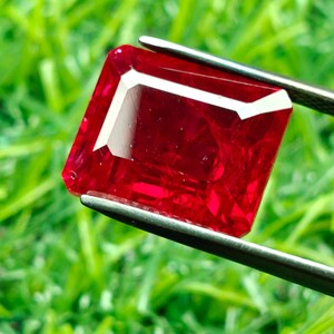 May include: A vibrant, rectangular-cut ruby gemstone held by tweezers against a blurred green background. The ruby is a deep, saturated red with white facets, showcasing its clarity and brilliance. The tweezers are silver-colored.