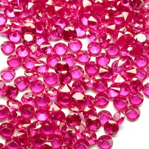 Może przedstawiać: Zbliżenie na stos małych, fasetowanych kamieni szlachetnych w kolorze rubinu. Kamienie są rozrzucone po całej ramie, odbijając światło i odbijając jasny, różowo-czerwony odcień. Kamienie szlachetne są prawdopodobnie używane do celów dekoracyjnych.