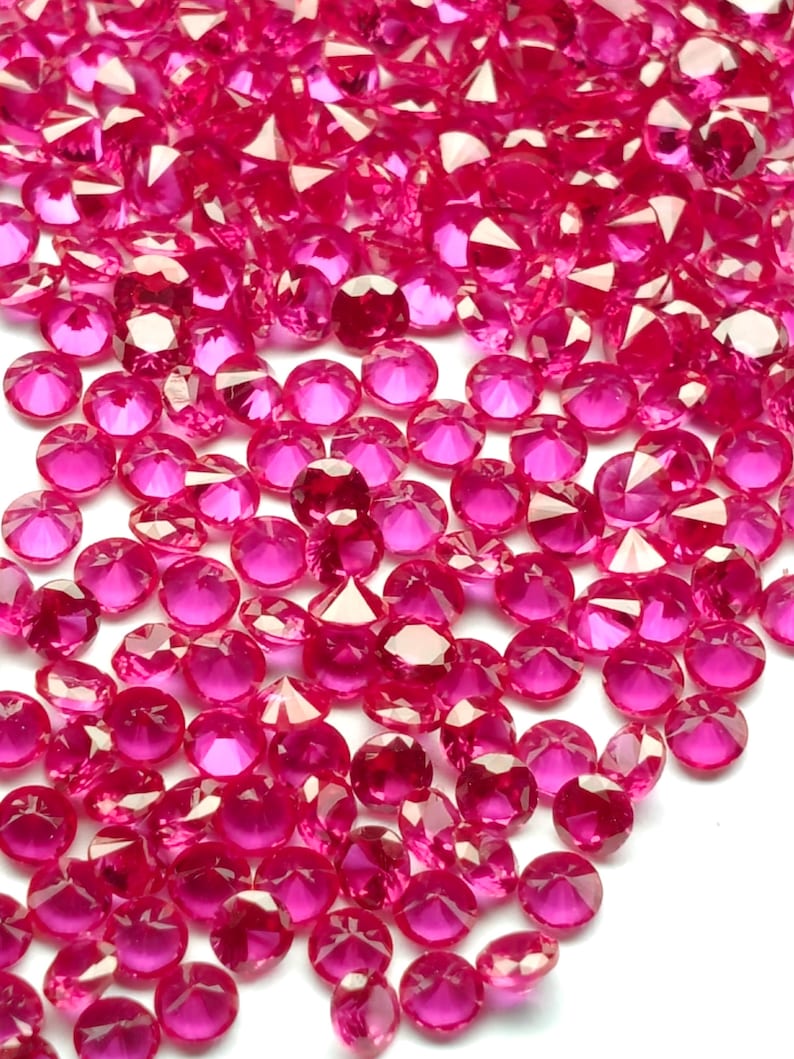 May include: A close-up shot of a pile of small, faceted, ruby-colored gemstones. The gems are scattered across the frame, catching the light and reflecting a bright, pinkish-red hue. The gems are likely used for decorative purposes.