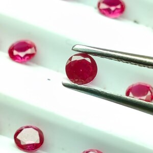 May include: A collection of faceted, oval-shaped rubies in varying sizes. One ruby is held by tweezers, showcasing its deep red color and clarity. The gems are displayed on a white surface, highlighting their brilliance.