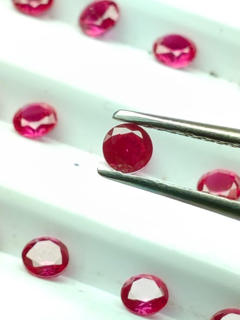 May include: A collection of faceted, oval-shaped rubies in varying sizes. One ruby is held by tweezers, showcasing its deep red color and clarity. The gems are displayed on a white surface, highlighting their brilliance.