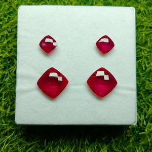 May include: Four faceted, cushion-cut, red gemstones are displayed on a white surface, set against a backdrop of green grass. Two smaller gems are above two larger gems. The gems have a vibrant, polished appearance.